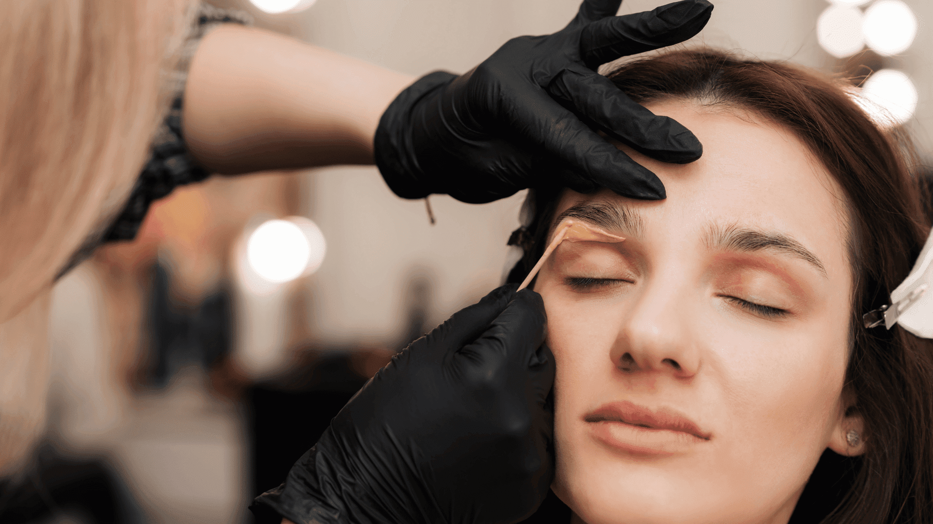 Woman getting eyebrows shaped by beautician wearing black gloves.