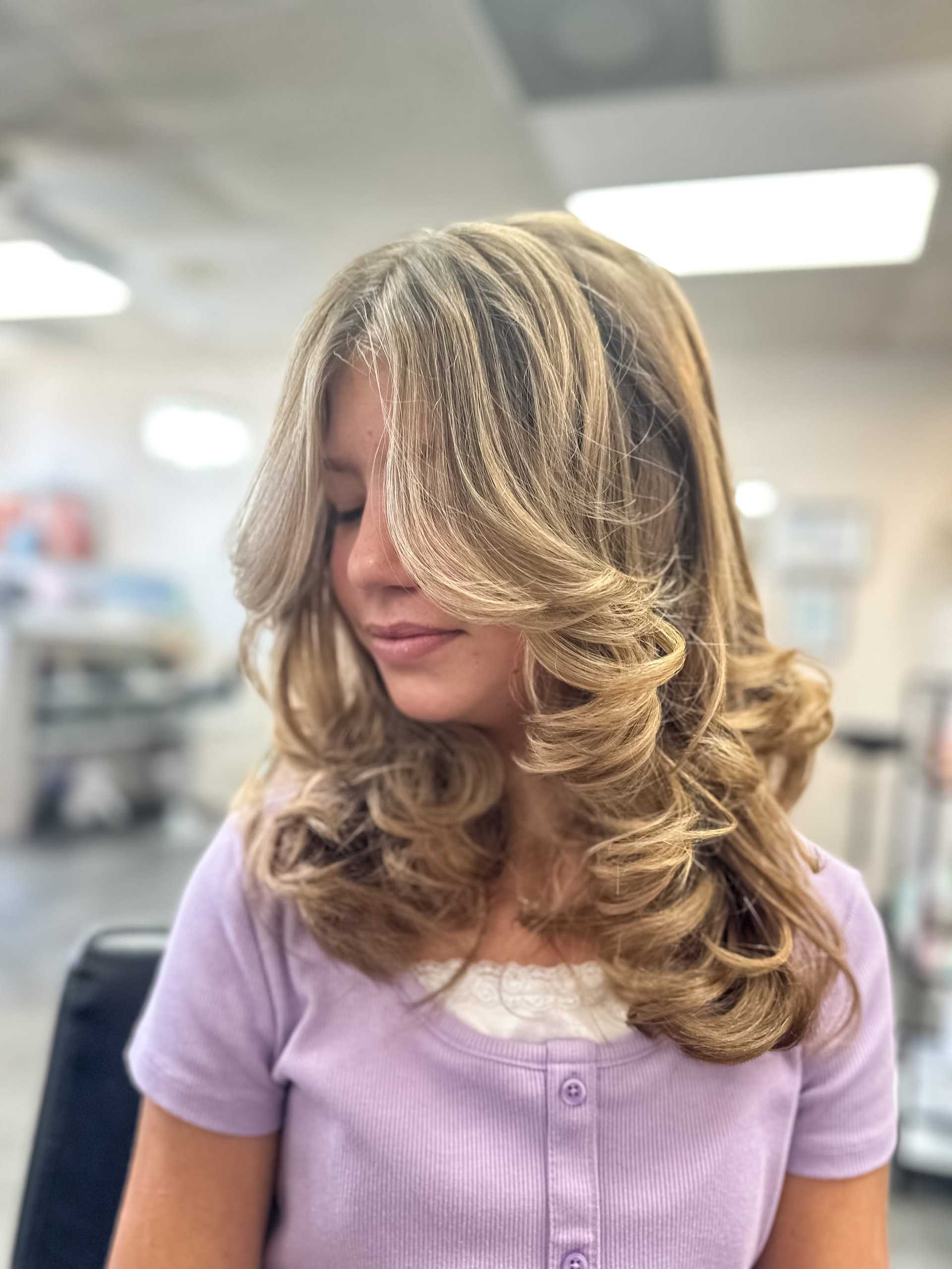 Woman with wavy, layered hairstyle in a salon, wearing a purple top.