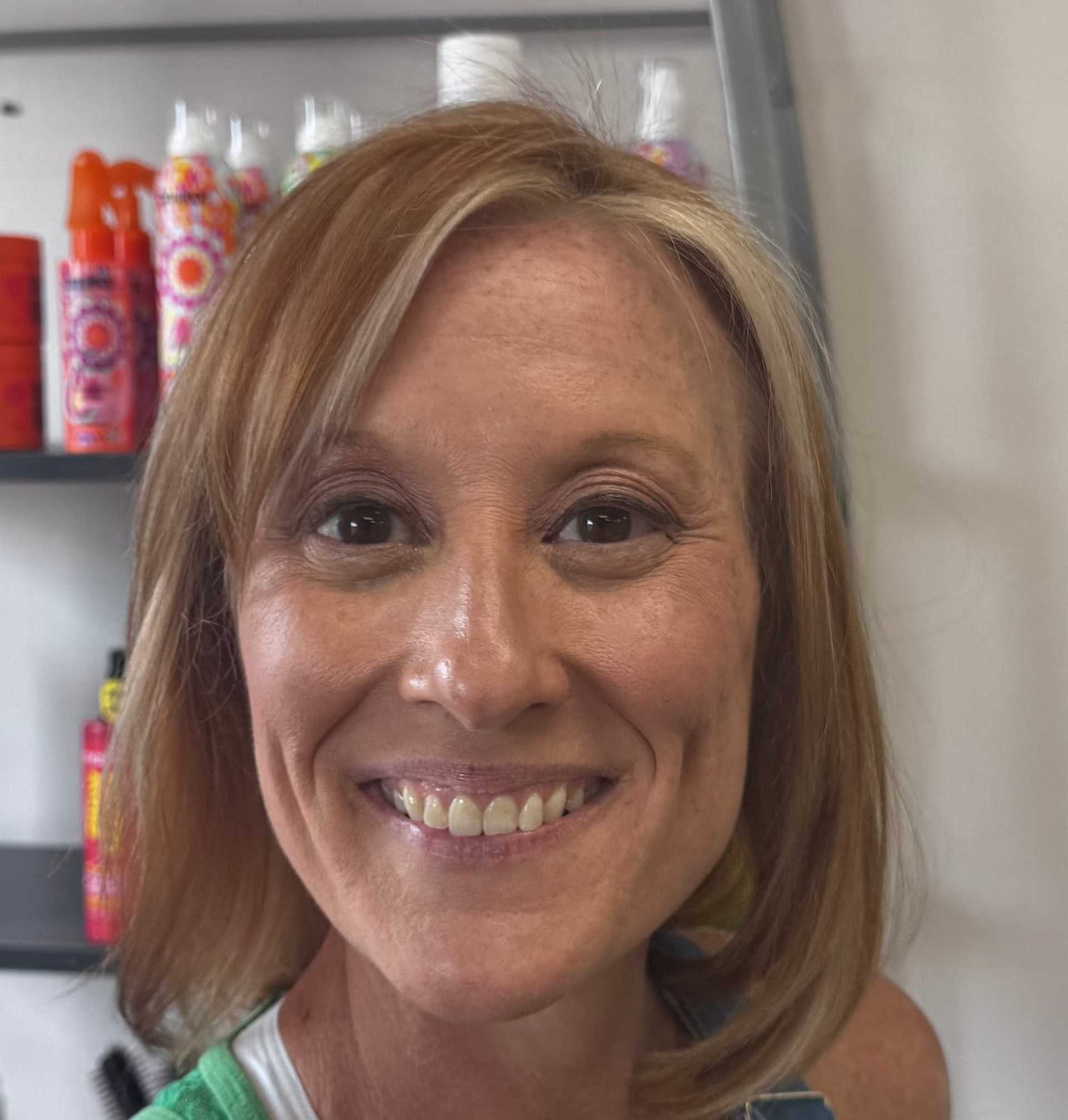 Smiling woman with light brown hair in front of haircare products on shelves.