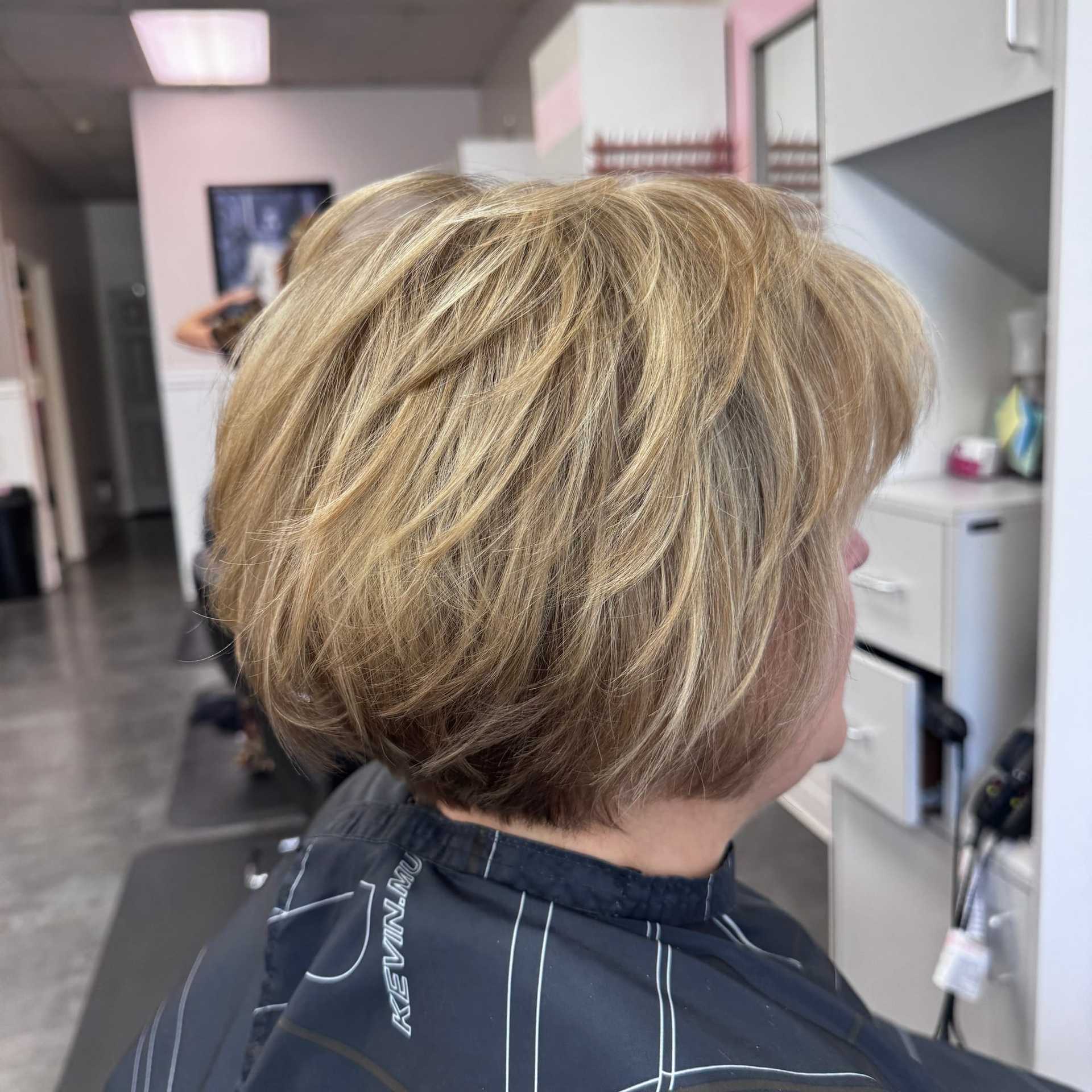 Short blonde bob haircut in a salon chair, stylist working in the background.