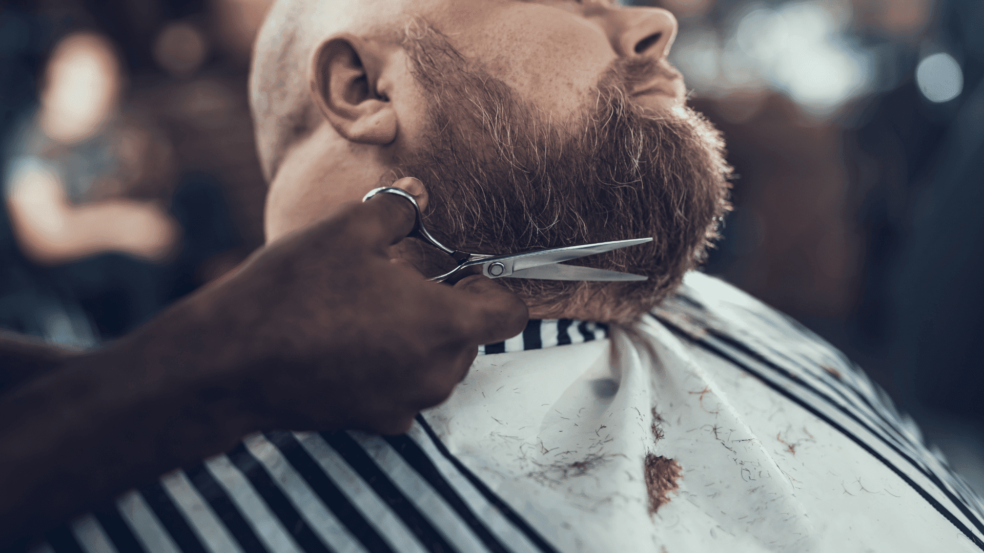 Bearded man getting a trim at a barbershop, wearing a striped cape.